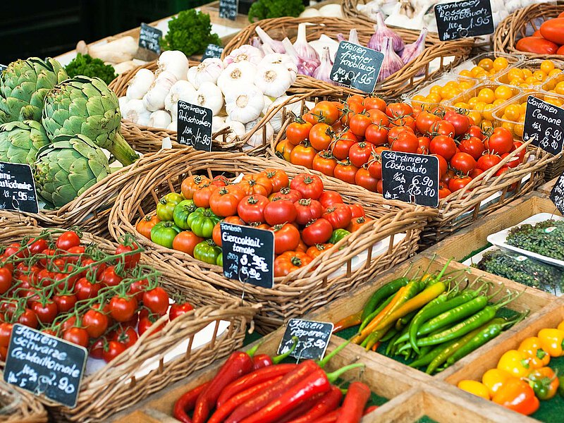 Market stall with baskets of tomatoes, artichokes, garlic, yellow cherry tomatoes, and peppers, each marked with small price signs.
