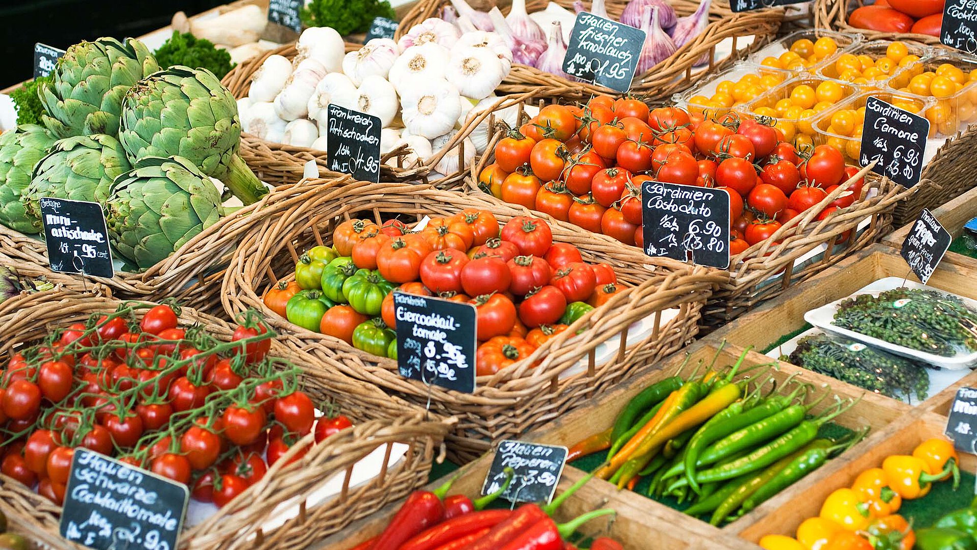 Market stall with baskets of tomatoes, artichokes, garlic, yellow cherry tomatoes, and peppers, each marked with small price signs.