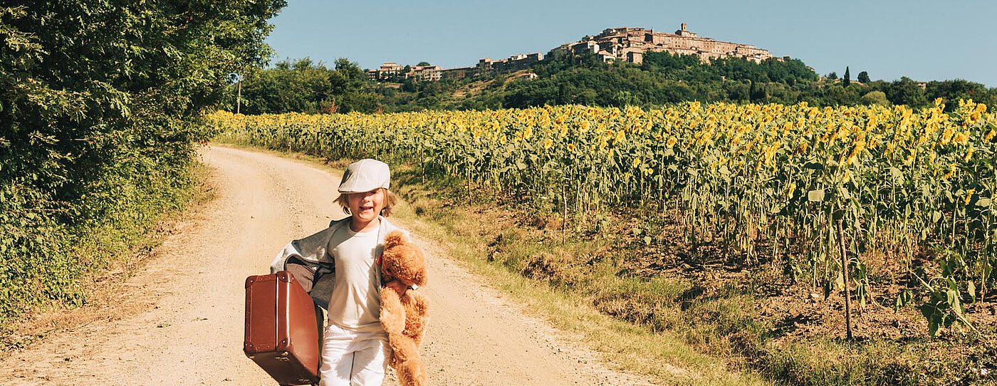 A child runs along a dirt road, carrying a suitcase and a teddy bear. The scene includes vibrant sunflowers on either side, with a scenic village in the distance under a clear blue sky.