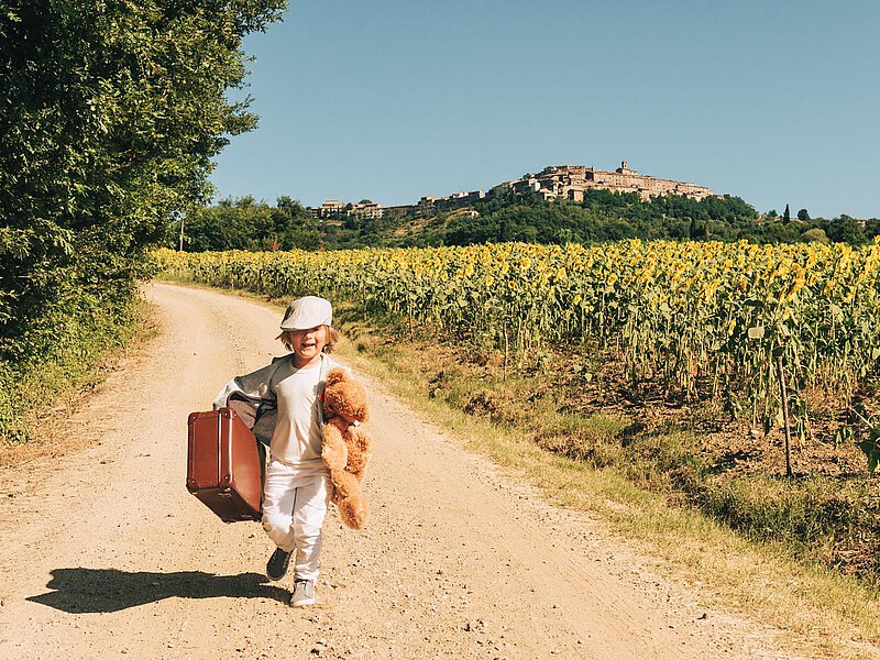 A child runs along a dirt road, carrying a suitcase and a teddy bear. The scene includes vibrant sunflowers on either side, with a scenic village in the distance under a clear blue sky.