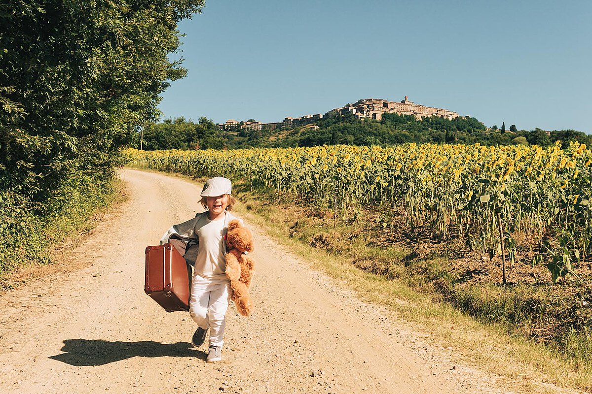 A child runs along a dirt road, carrying a suitcase and a teddy bear. The scene includes vibrant sunflowers on either side, with a scenic village in the distance under a clear blue sky.