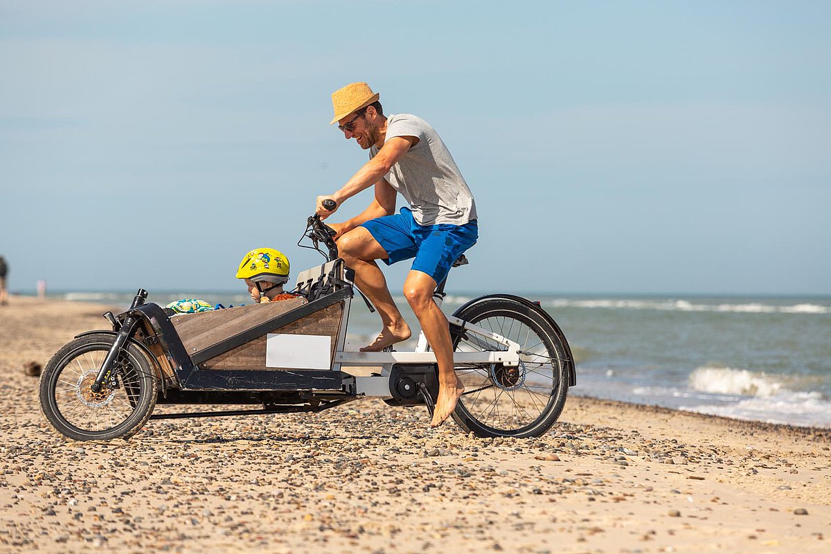 A man pedaling a cargo bike on the beach, with a child sitting in the cargo area, surrounded by sand and ocean waves under a sunny sky.