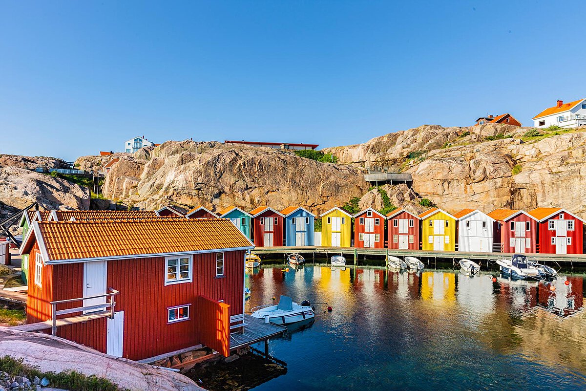 Bunte Holzhäuser entlang eines Hafens an Schwedens Westküste, mit Reflexionen im Wasser und felsigen Klippen im Hintergrund, die die Schönheit der Küstenlandschaft präsentieren.