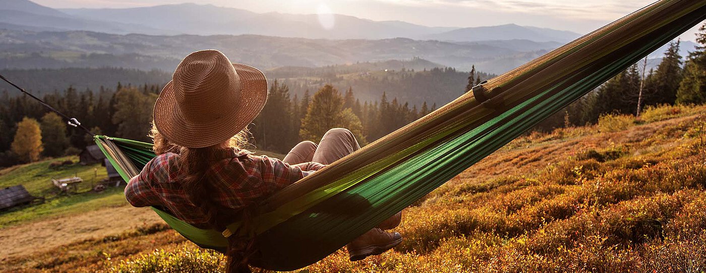 Frau mit Hut entspannt in einer Hängematte und blickt über eine herbstliche Berglandschaft.