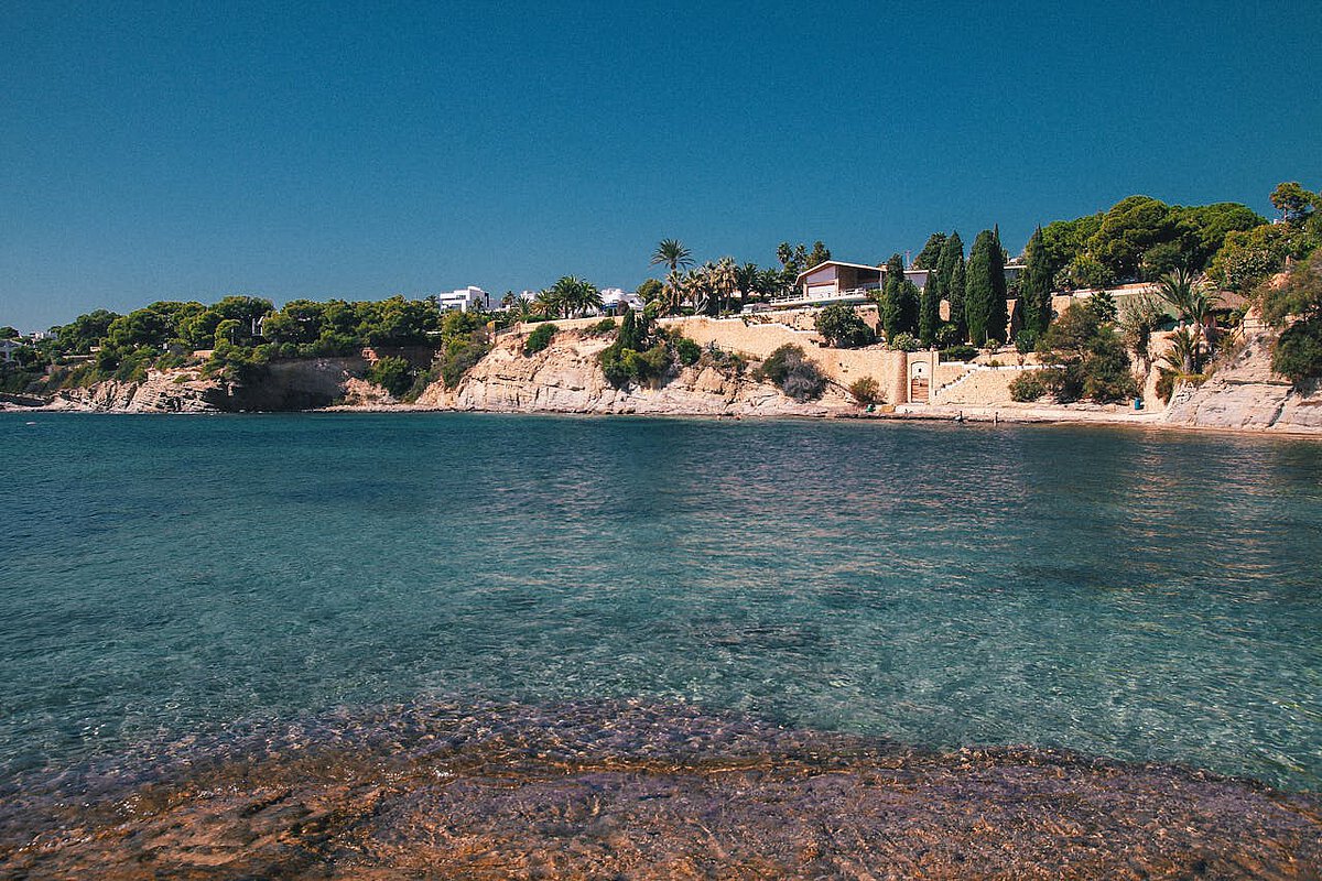 Crystal clear sea on the Costa Blanca in the province of Valencia, a holiday home by the sea and Mediterranean vegetation in the distance.