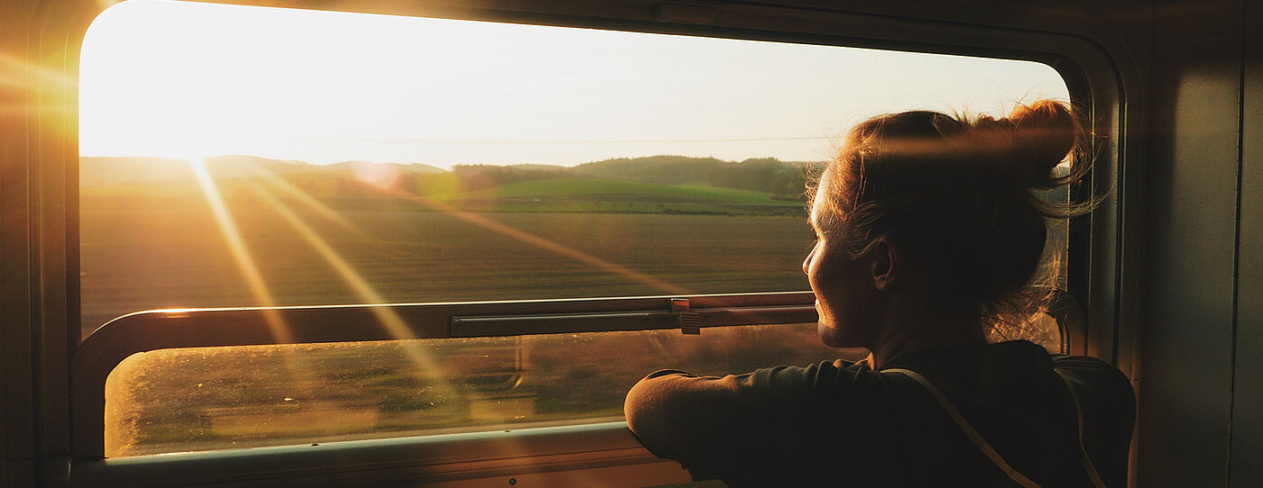 Eine Person lehnt am offenen Zugfenster und blickt in den Sonnenuntergang über weiten Feldern, während goldenes Licht den Innenraum erhellt.
