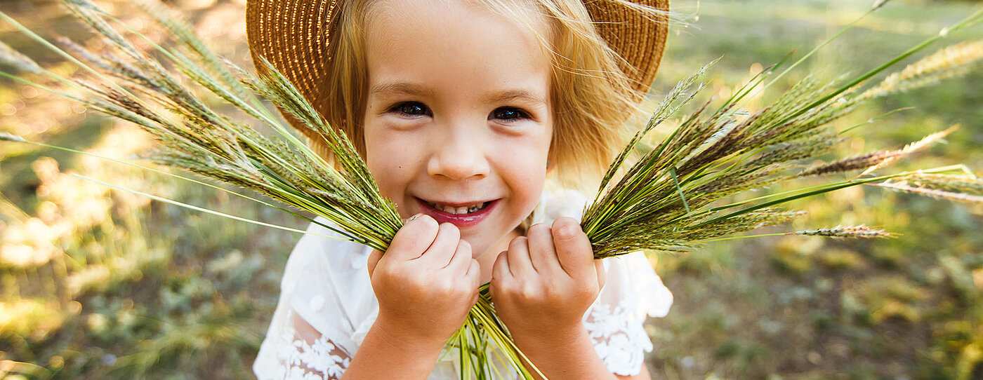 Lächelndes Kind mit Strohhut hält ein Büschel langer Gräser in beiden Händen. Hintergrund mit Wiese und Bäumen im warmen Licht.
