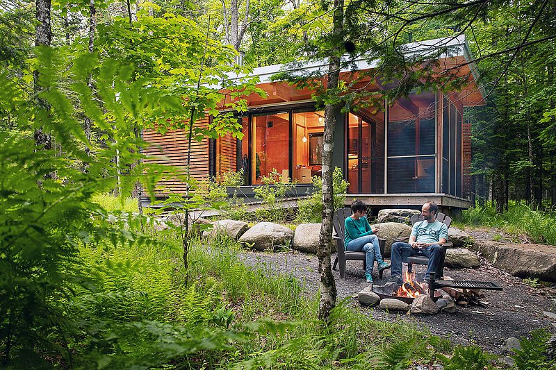 Campfire Moments in a Romanian Woodland Cabin A couple relaxes in wooden chairs by a crackling campfire, surrounded by lush forest greenery. Behind them, a modern cabin with large glass windows glows warmly from within. Ferns and moss cover the forest floor, creating a peaceful, secluded atmosphere close to nature.