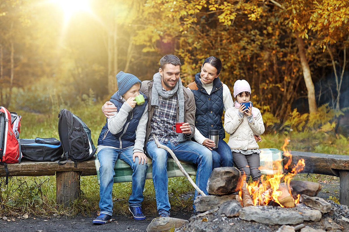 A family of four sitting around a campfire in the woods, enjoying warm drinks. They are surrounded by autumn leaves and a cozy atmosphere.