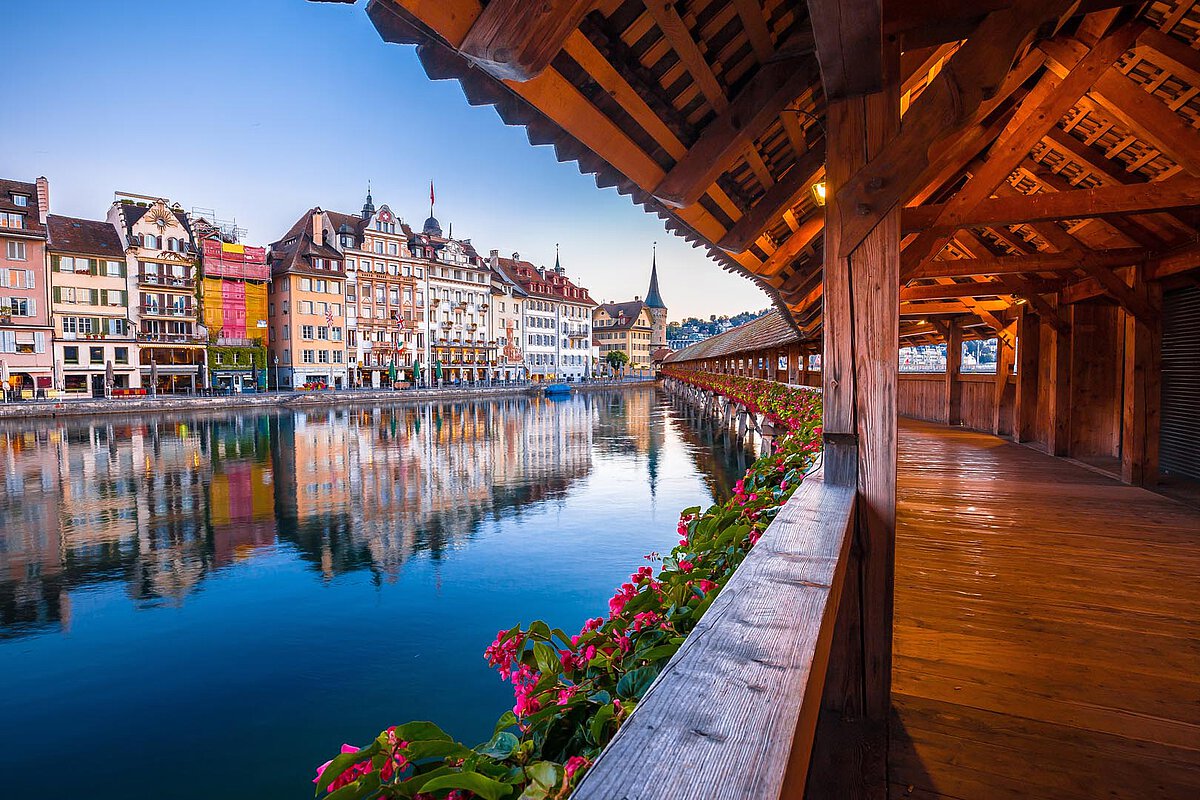Blick auf den Vierwaldstättersee mit bunten Gebäuden, die sich im Wasser spiegeln, und einem Holzsteg mit Blumen im Vordergrund.