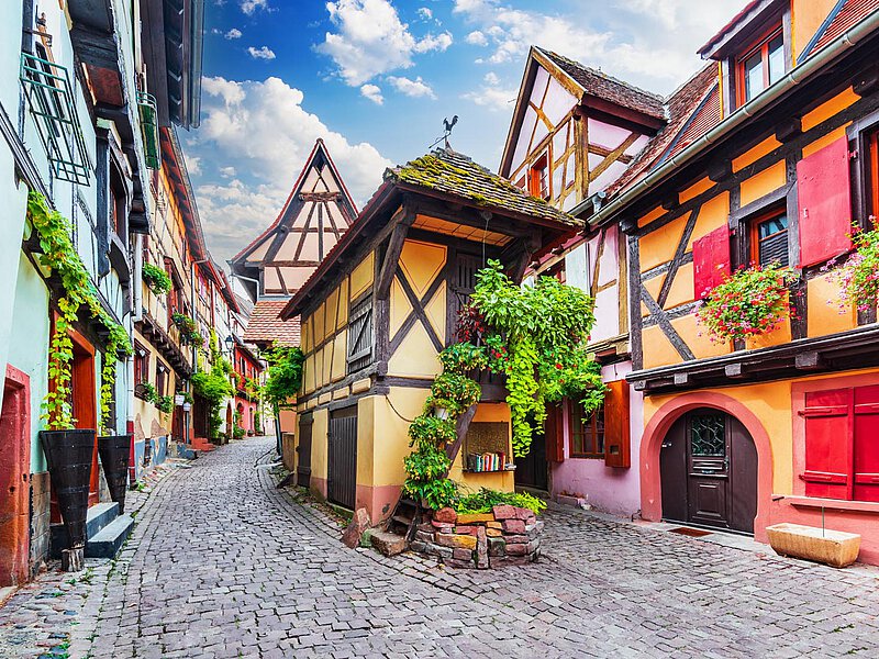 Idyllische Gasse in Eguisheim mit Fachwerkhäusern, Kopfsteinpflasterstraßen und bunten Blumenkästen unter einem teilweise bewölkten Himmel.