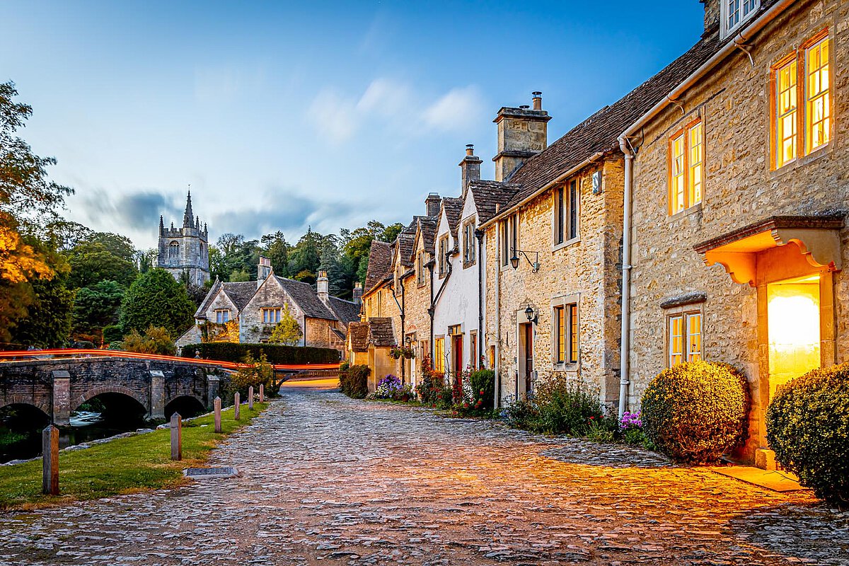 Charming village in Great Britain at dusk, with stone houses, a cobblestone path, and a historic bridge illuminated by warm window lights.