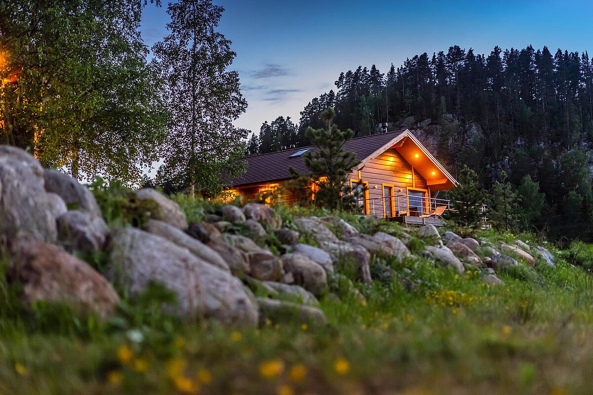 Wooden cabin illuminated at dusk, surrounded by rocks and trees, set in a serene landscape in Slovenia.