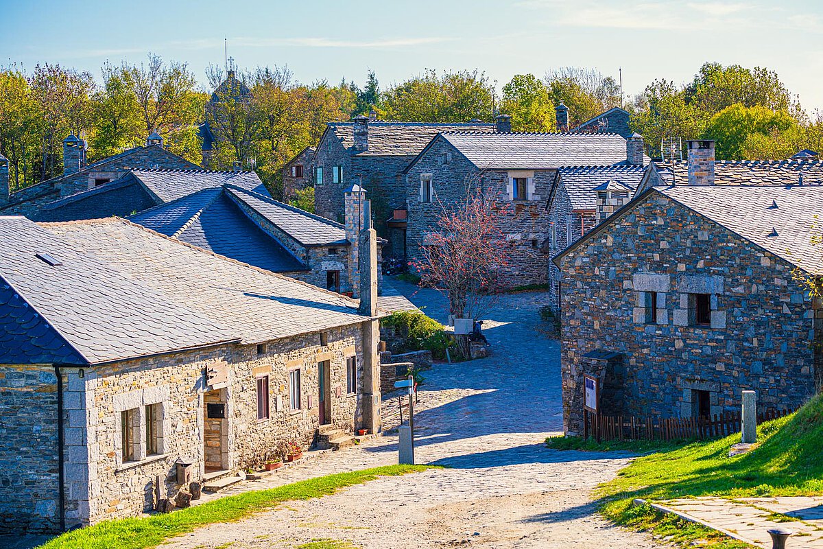 Traditional Galician village with stone houses and slate roofs along a cobblestone path, set amidst greenery and trees under a clear sky.