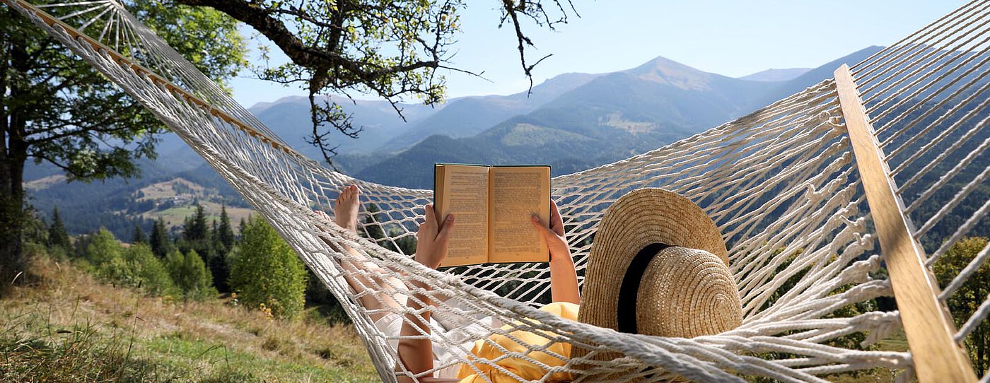 A person lying in a hammock, holding a book, with mountains in the background. They are wearing a wide-brimmed hat, and sunlight filters through the trees, creating a tranquil outdoor setting.