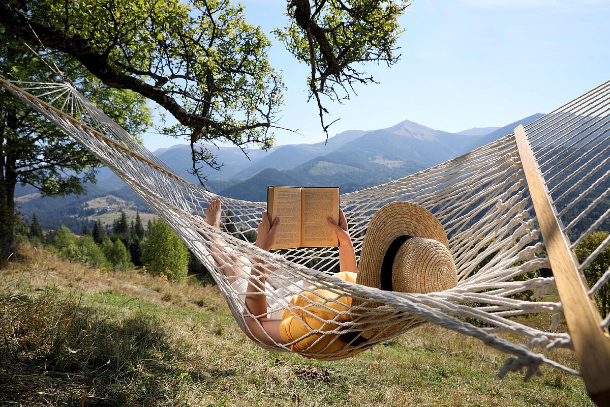 A person lying in a hammock, holding a book, with mountains in the background. They are wearing a wide-brimmed hat, and sunlight filters through the trees, creating a tranquil outdoor setting.