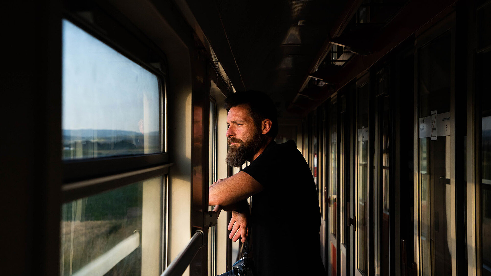A man stands in the corridor of a train, looking out of the window at the passing landscape in warm evening light.