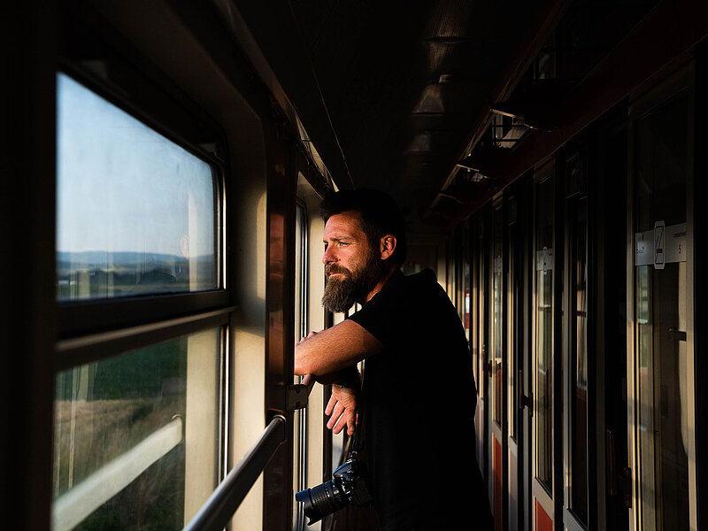 A man stands in the corridor of a train, looking out of the window at the passing landscape in warm evening light.