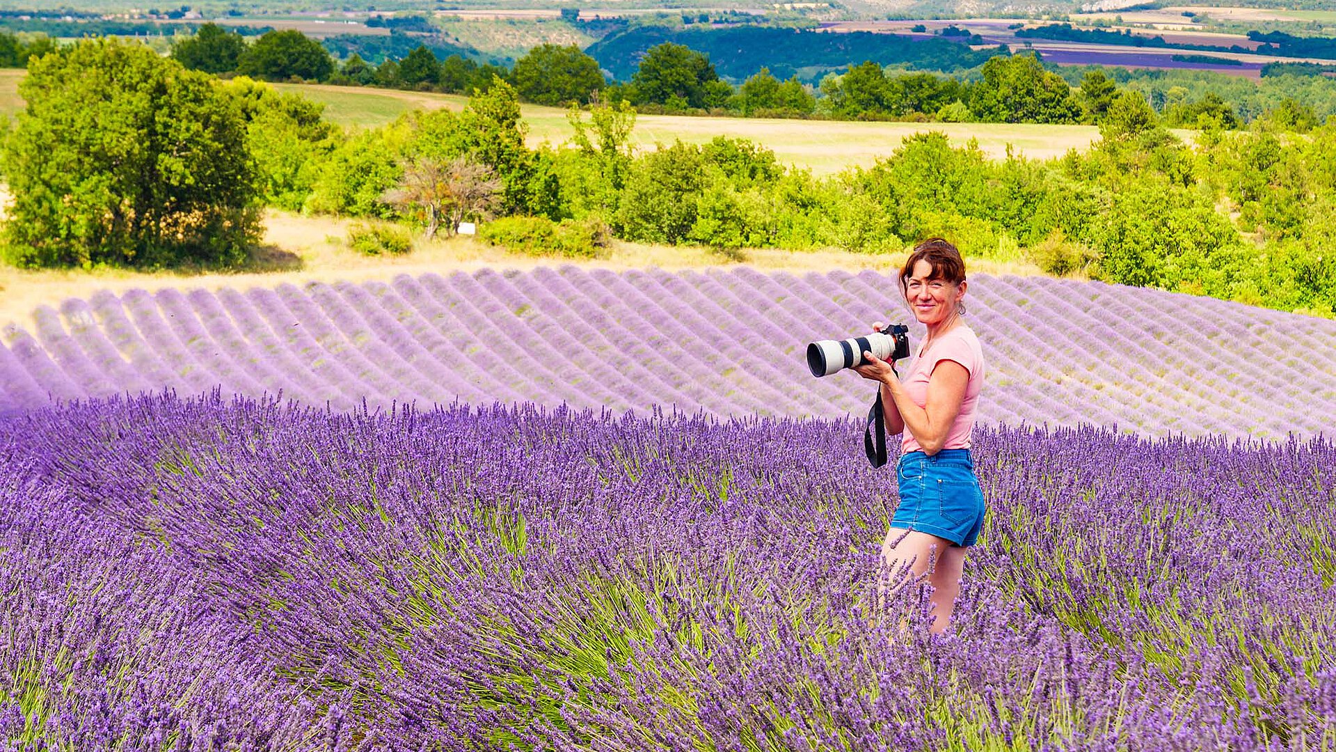 Frau mit Kamera steht in einem blühenden Lavendelfeld. Im Hintergrund sind grüne Hügel und Berge zu sehen.