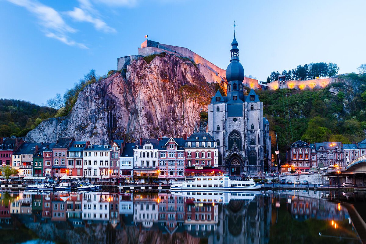 Flussuferansicht von Dinant, Belgien, mit der Kollegiatkirche Notre Dame und einer Festung an der Klippe bei Dämmerung, mit Spiegelungen im Wasser.