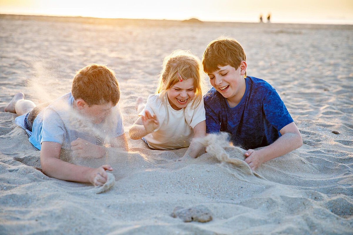 Three children playing in the sand on a beach, laughing and throwing sand as the sun sets in the background.
