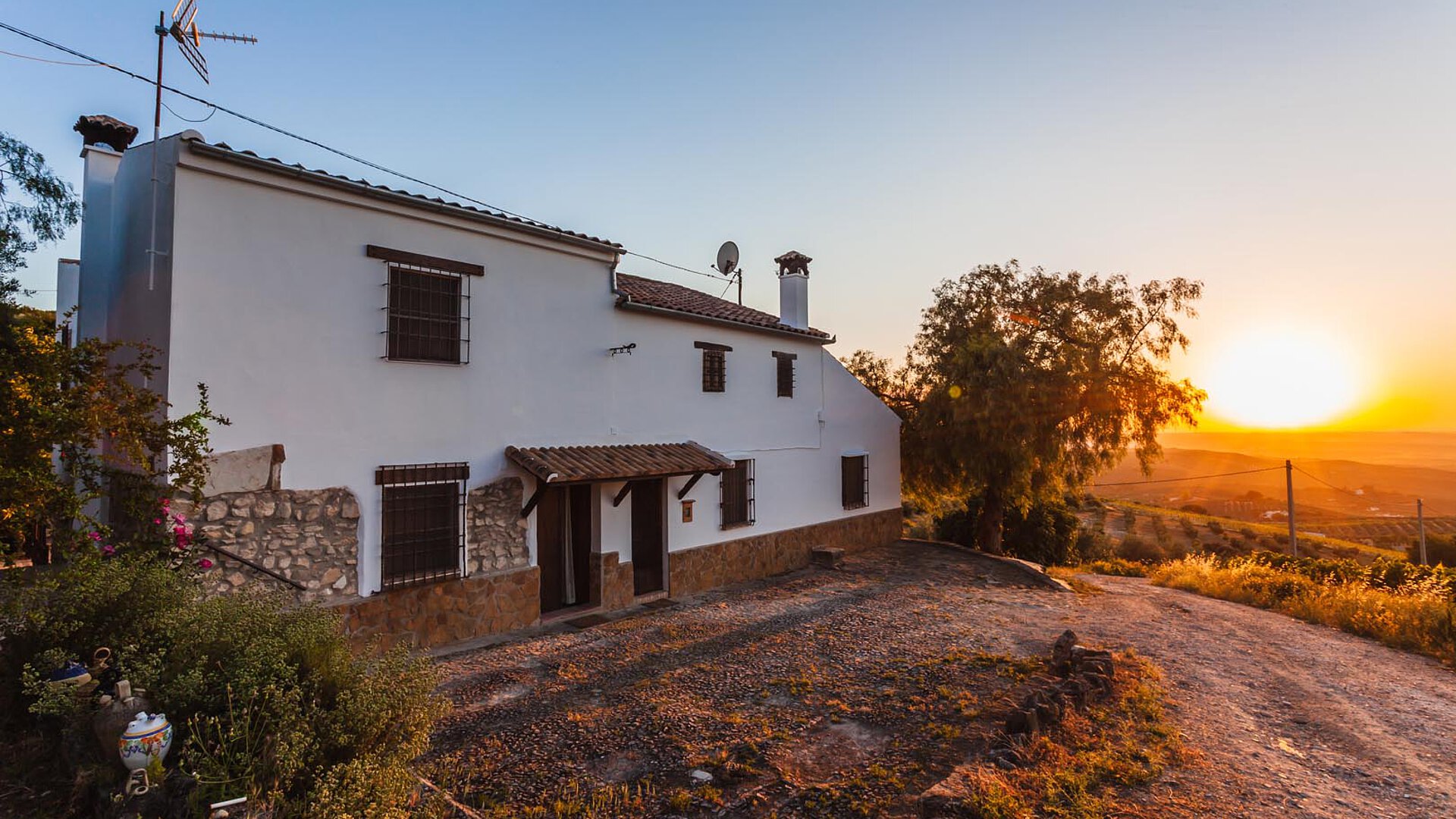 White countryside house with stone accents at sunset, surrounded by gardens and a gravel path, creating a peaceful rural scene.