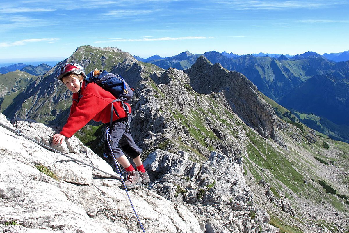 Ein Kind in einem roten Pullover klettert an einem felsigen Hang in den Bergen, gesichert durch ein Seil. Die Landschaft zeigt zerklüftete Gipfel und grüne Täler unter einem strahlend blauen Himmel und betont den Nervenkitzel des Kletterns im Freien.