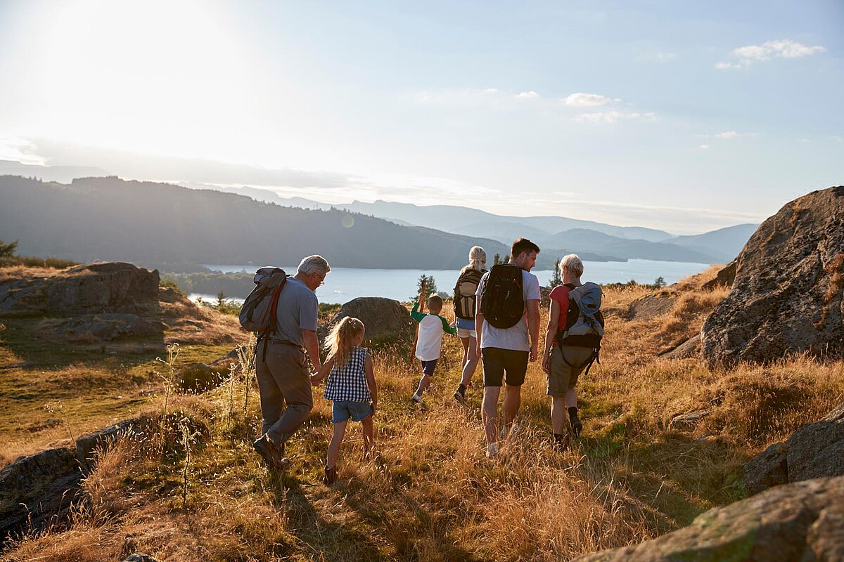 A family of six hiking on a trail with backpacks, surrounded by hills and a lake in the background during sunset.