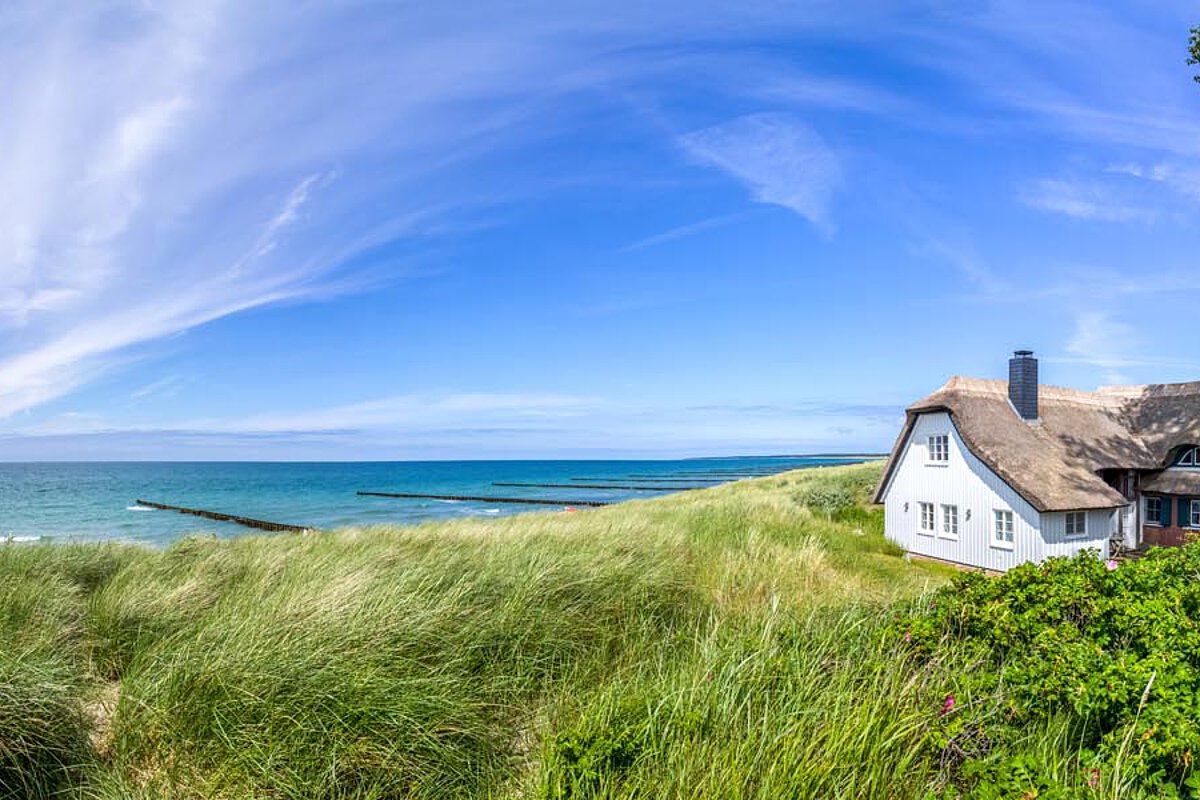 Panoramic view of the Baltic Sea coast with dunes, grass, and a thatched-roof house under a blue sky.