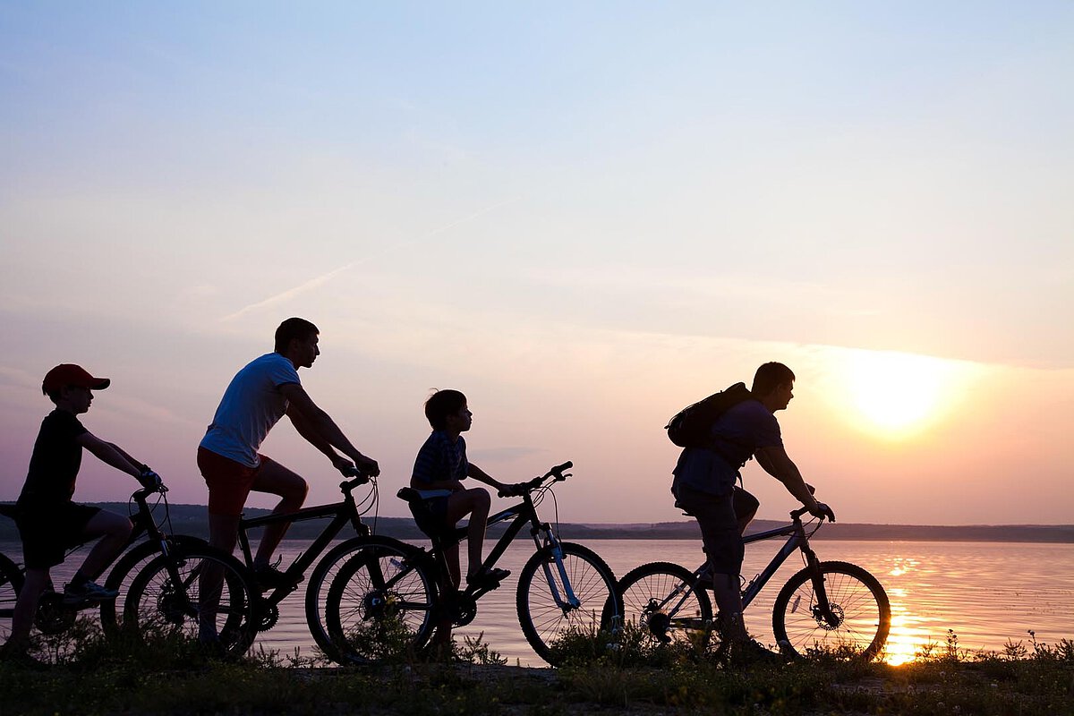 Four people ride bicycles along a lakeside path at sunset, with the sun setting behind them and reflecting on the water.