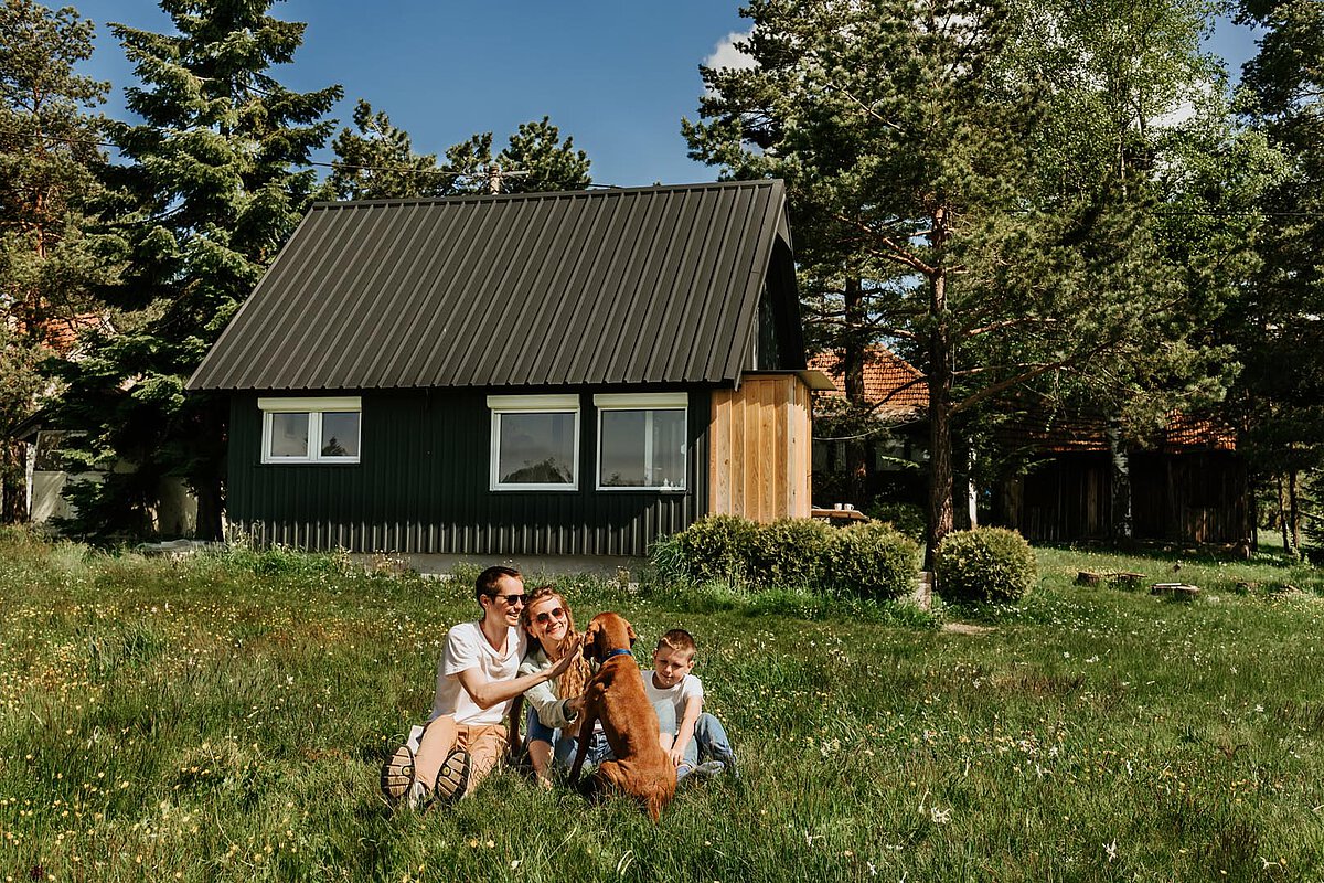 Eine Familie sitzt im Gras nahe ihrem Ferienhaus und spielt mit einem Hund. Die Mutter und das Kind beschäftigen sich mit dem Haustier, während das Ferienhaus und die Bäume eine charmante Kulisse in einer sonnigen Umgebung bilden.
