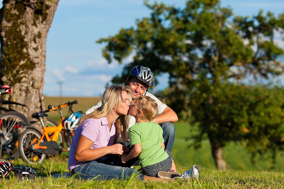 A mother and father share a kiss with their young child on a grassy area, with bicycles in the background and a clear blue sky above.