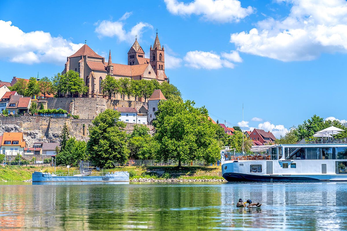 Historic church on a hillside in Baden-Württemberg, a boat on the river, and ducks swimming, with a bright blue sky.