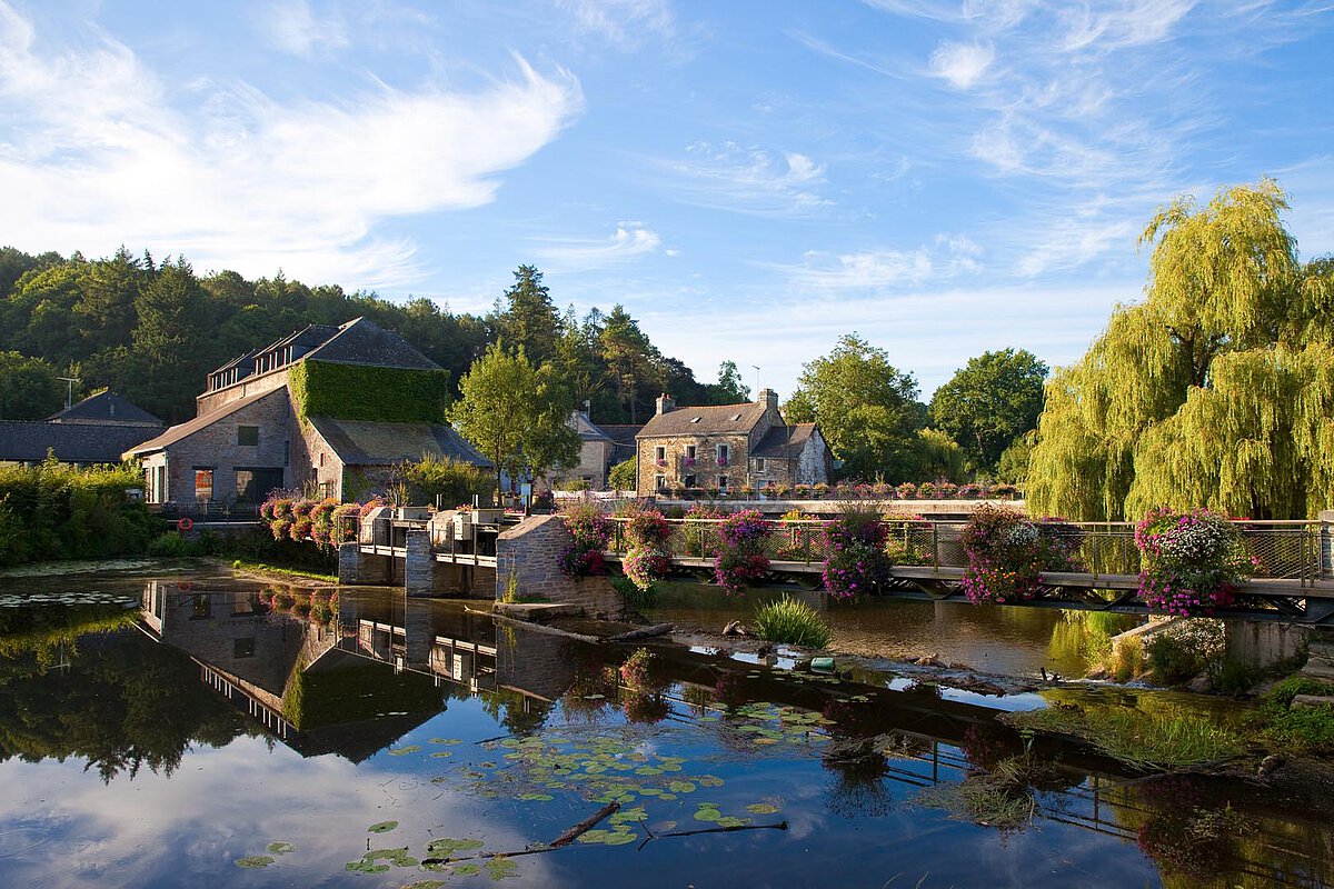 Country estate with buildings and flowers reflected in calm water, under a blue sky.