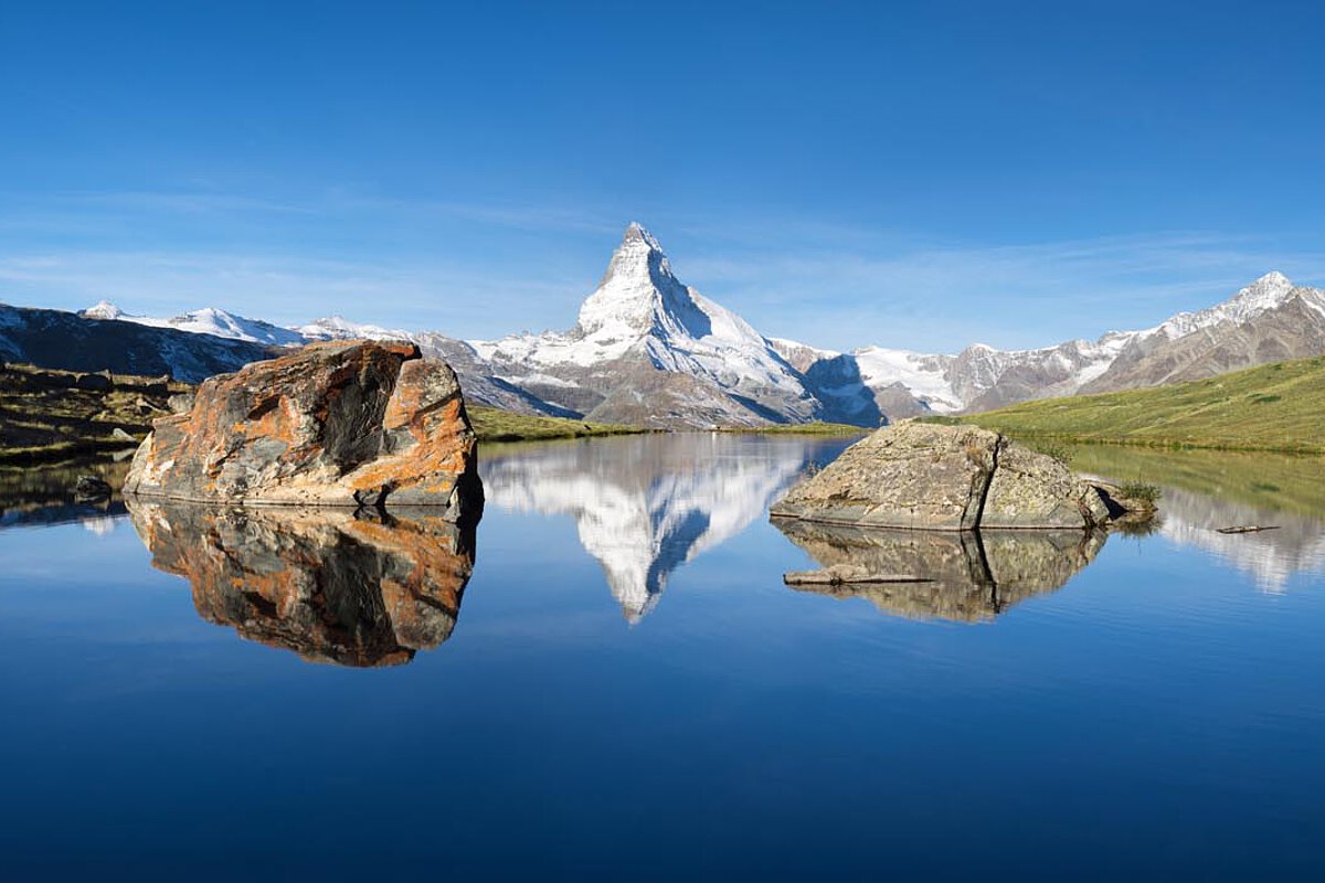 Reflexion des Matterhorns in einem See, umgeben von Bergen und grünen Hügeln unter einem klaren blauen Himmel.