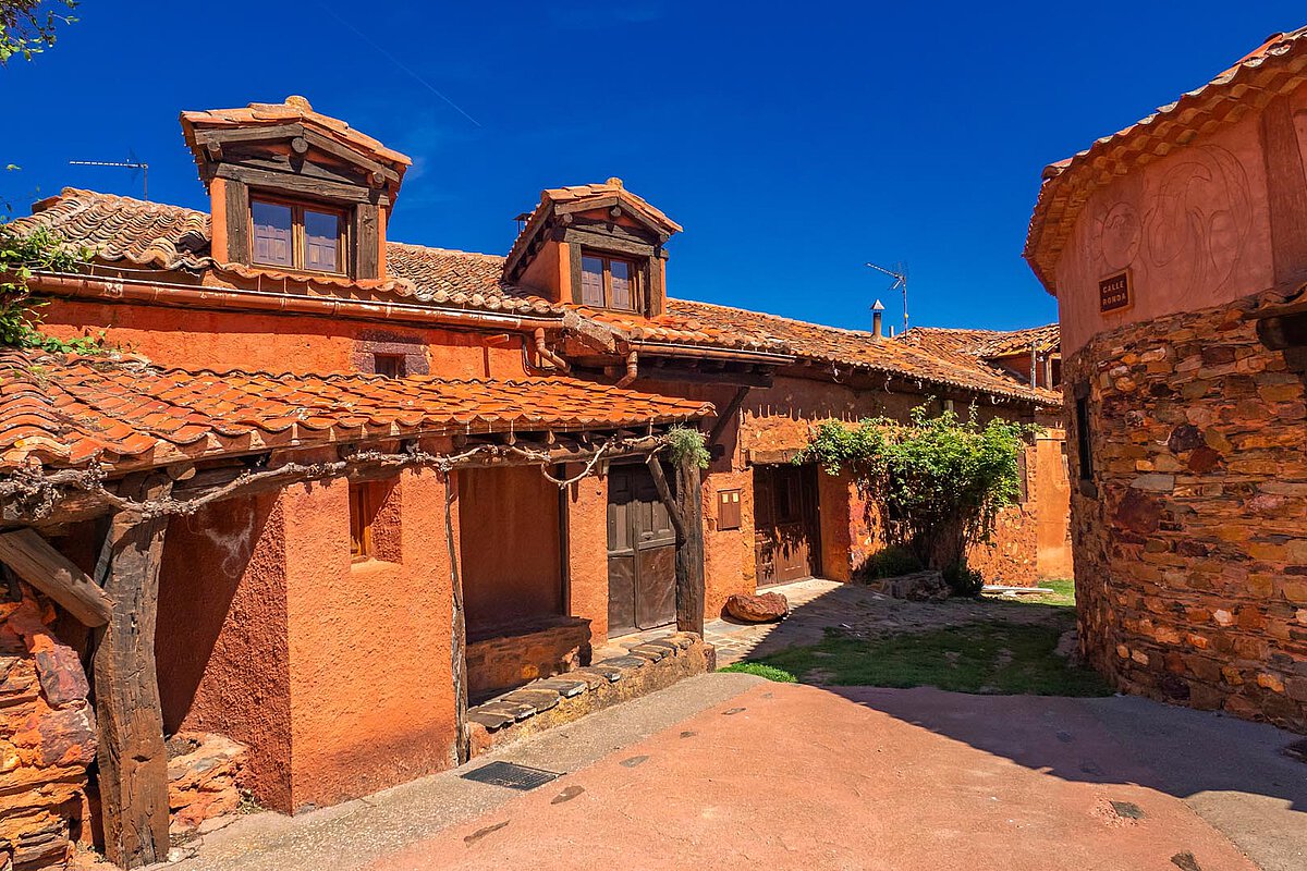 Charming rural scene in Spain with terracotta-roofed buildings and a narrow pathway, surrounded by greenery and under a clear blue sky.
