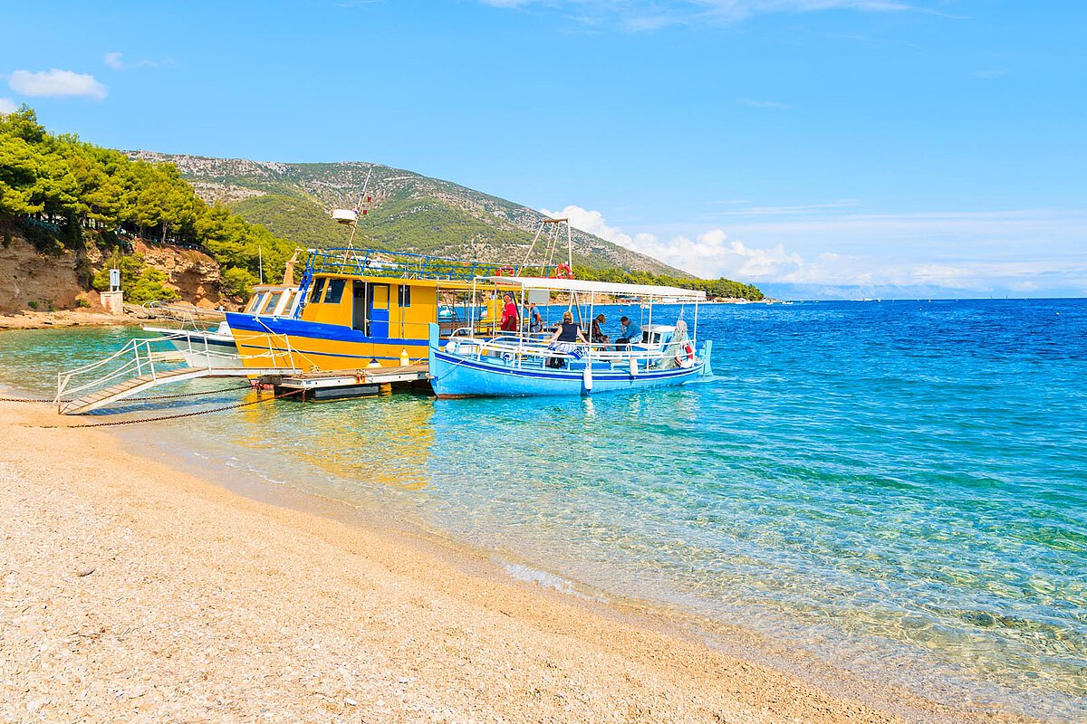 Bunte Fischboote, die an einem sandigen Strand in Portorož festgemacht sind, mit klarem türkisfarbenem Wasser und grünen Hügeln im Hintergrund an einem sonnigen Tag.