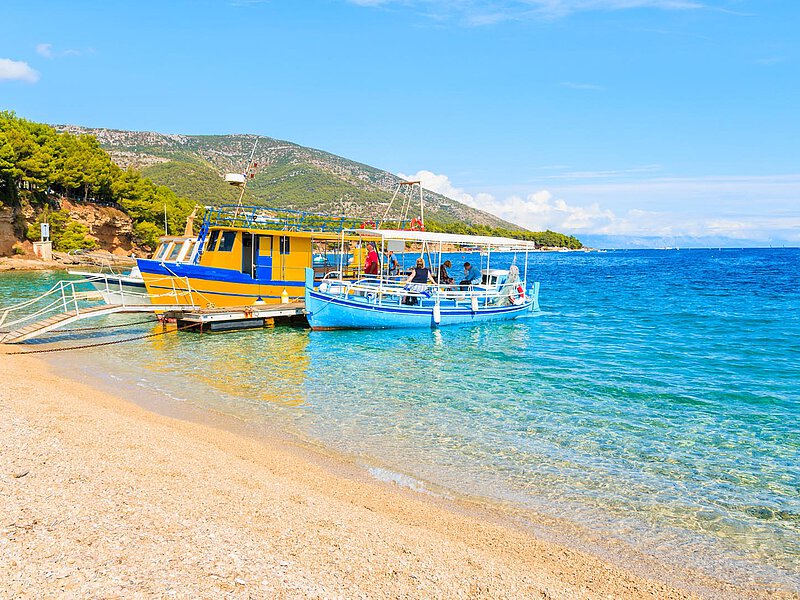 Bunte Fischboote, die an einem sandigen Strand in Portorož festgemacht sind, mit klarem türkisfarbenem Wasser und grünen Hügeln im Hintergrund an einem sonnigen Tag.