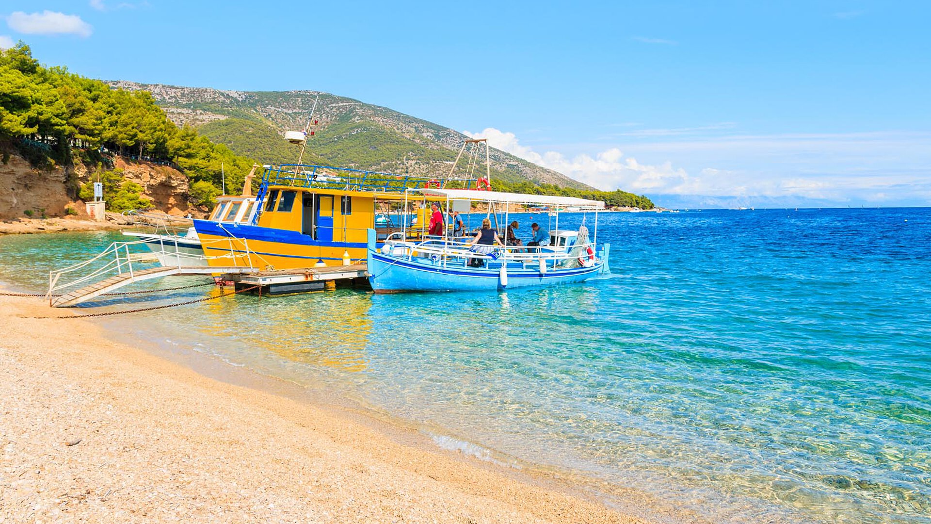 Bunte Fischboote, die an einem sandigen Strand in Portorož festgemacht sind, mit klarem türkisfarbenem Wasser und grünen Hügeln im Hintergrund an einem sonnigen Tag.